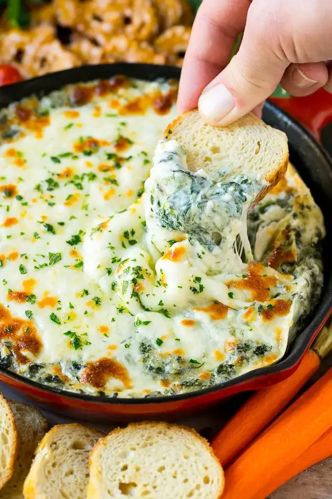 A piece of bread scooping into a pan of spinach dip.